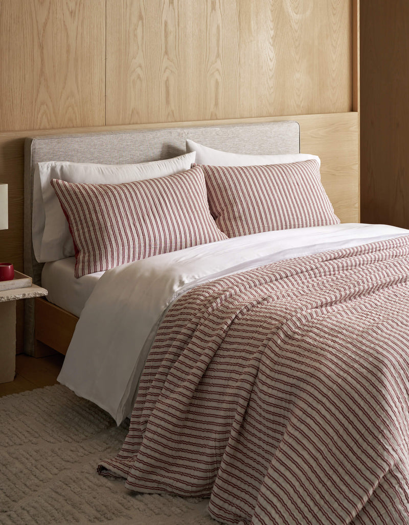 A neatly made bed features the Cozy Earth Waterfront Coverlet Bundle, white sheets, and a striped comforter with matching pillows, set against a light wood headboard and wall, complemented by a beige rug and a small bedside table.