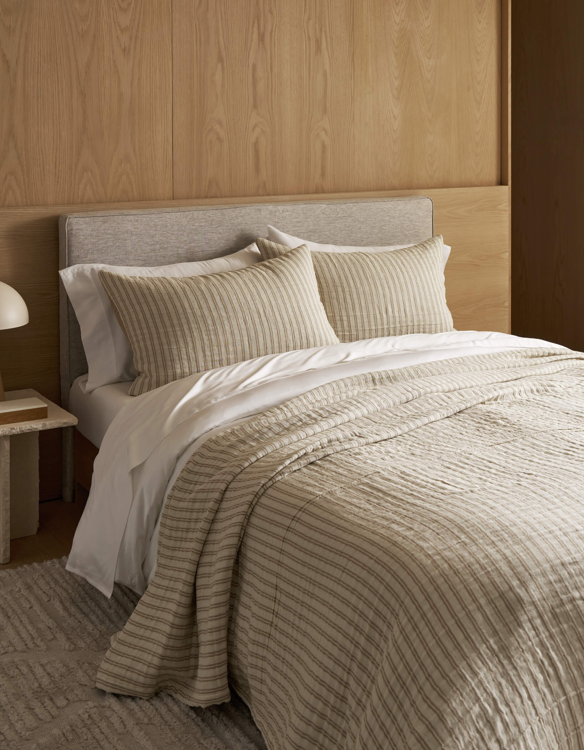 The cozy, minimalist bedroom features a Waterfront Coverlet Bundle by Cozy Earth on the bed with two pillows, set against a light wood panel wall. A small bedside table with a white lamp and a textured rug complete the scene.|Color:Sandalwood Stripe