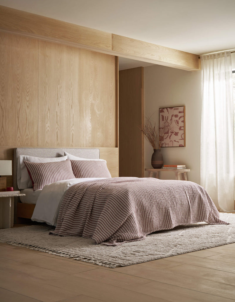 A modern bedroom featuring the Cozy Earth Waterfront Coverlet Bundle—pink-striped quilt and pillows—set against a light wood wall, with a beige rug, small side table, vase, framed abstract art, and sheer window curtains.
