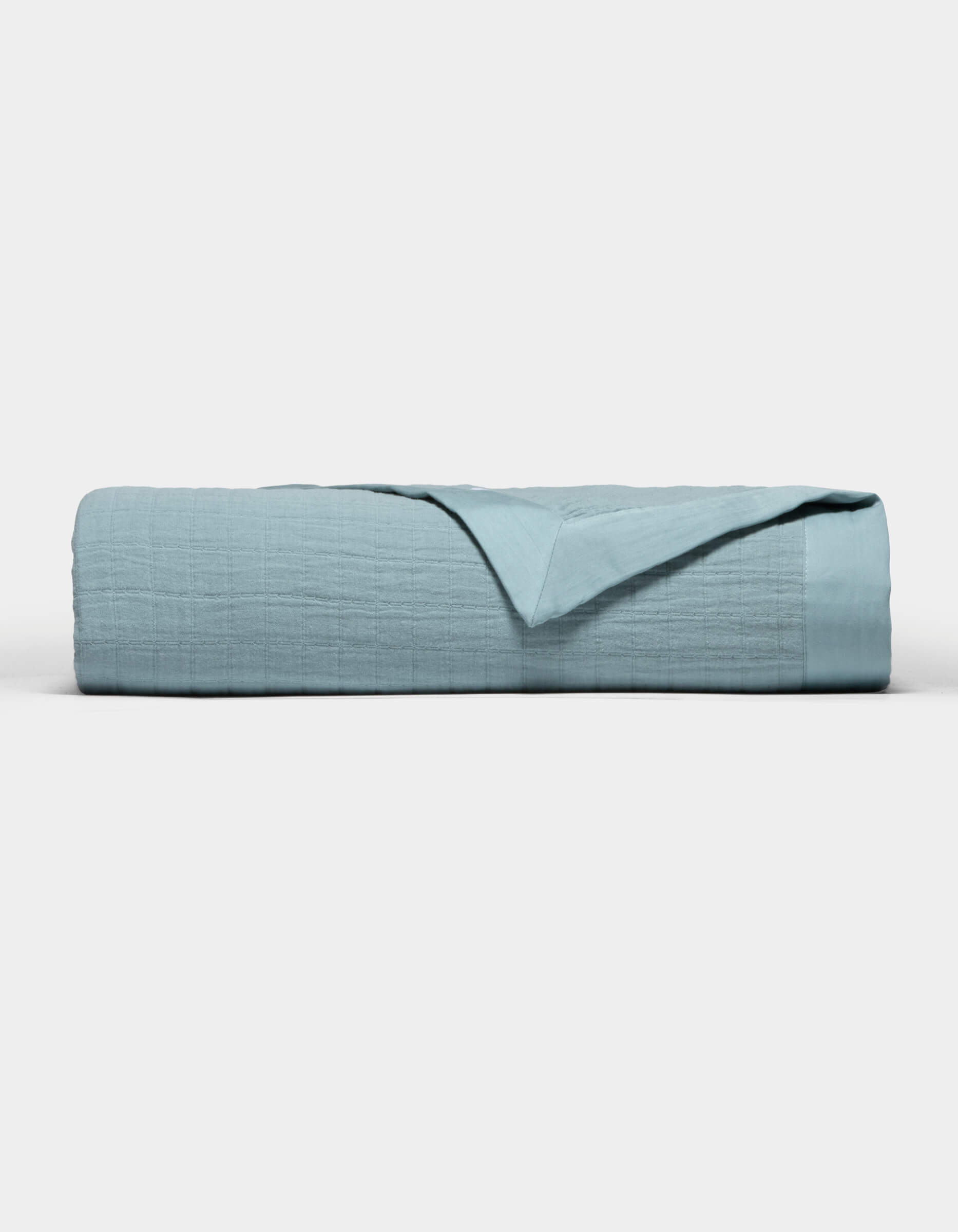 The Cozy Earth Waterfront Grid Matelasse Blanket in light blue is neatly folded against a plain white background, with the top layer turned back to show the inner fabric.|Color:River