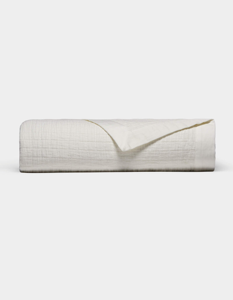 The Waterfront Grid Matelasse Blanket by Cozy Earth is shown neatly folded with a textured, crinkled surface against a plain light background. One corner is flipped over to reveal the underside.
