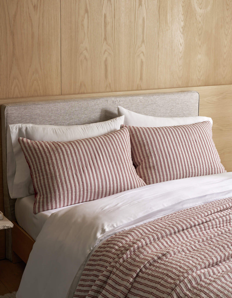 A Waterfront Coverlet Bundle by Cozy Earth, featuring white sheets, two pillows, and a comforter with red and white vertical stripes, is arranged on a bed with a light wood-paneled wall and a light gray upholstered headboard.