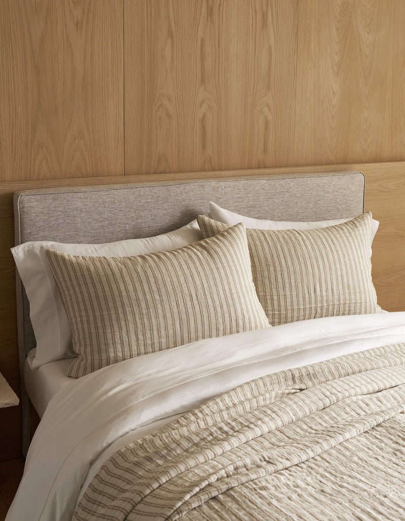 A neatly made bed features the Cozy Earth Waterfront Coverlet Bundle—beige upholstered headboard, white sheets, and cream ribbed pillows and comforter—set against a light wood-paneled wall.