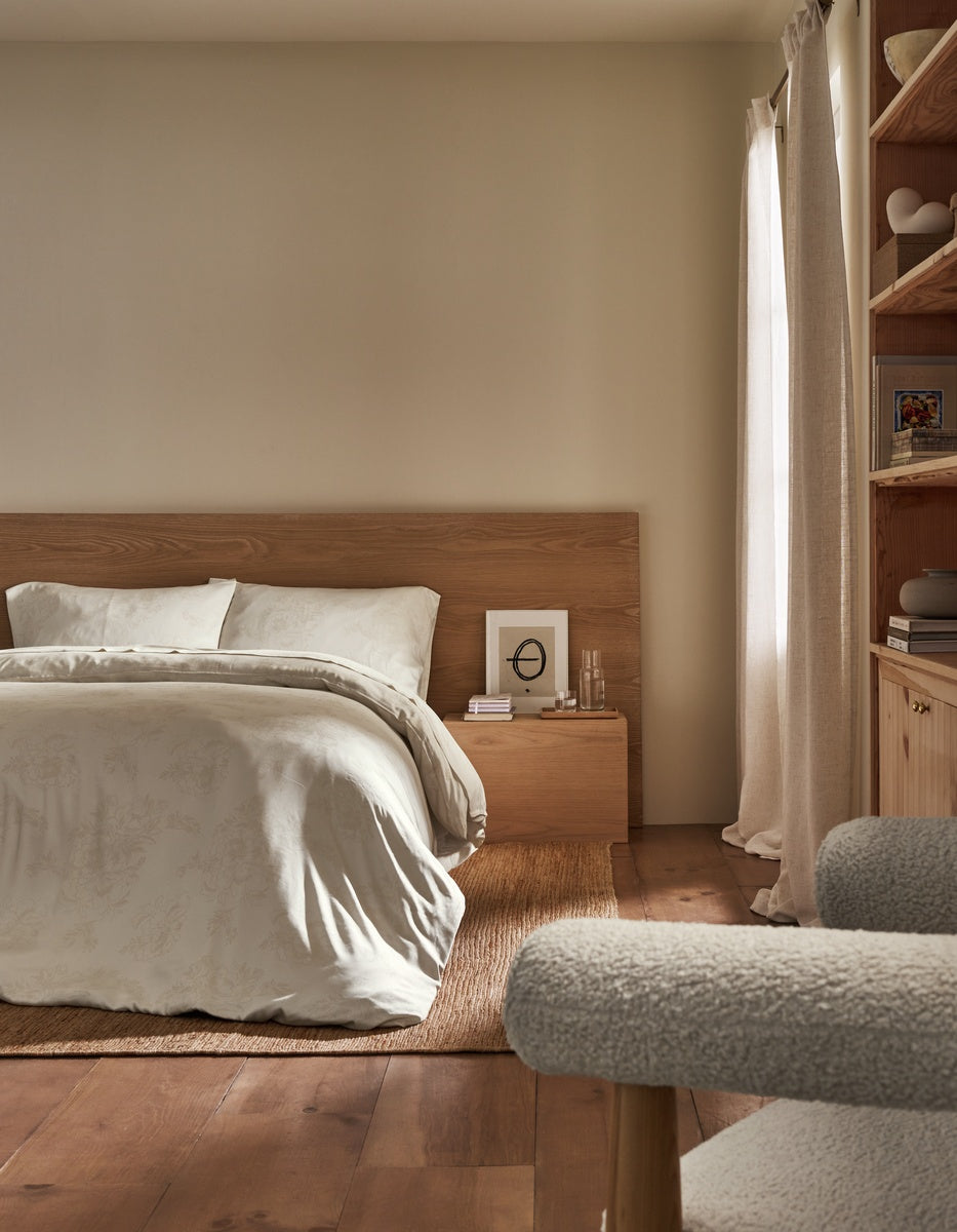 A minimalist bedroom scene featuring a bed with white bedding and a light wood headboard, styled in a warm neutral room with a wooden floor, beige walls, and a window with cream curtains.|Color:Sonoma Floral in Vintage White