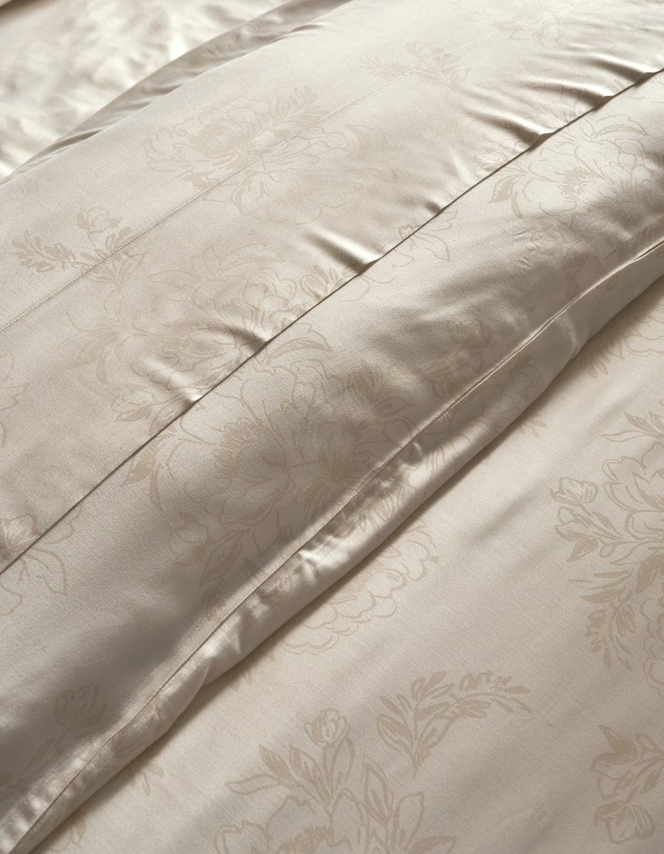 Close-up of a cream-colored satin duvet cover with a subtle floral damask pattern and stitched channel tufting. 