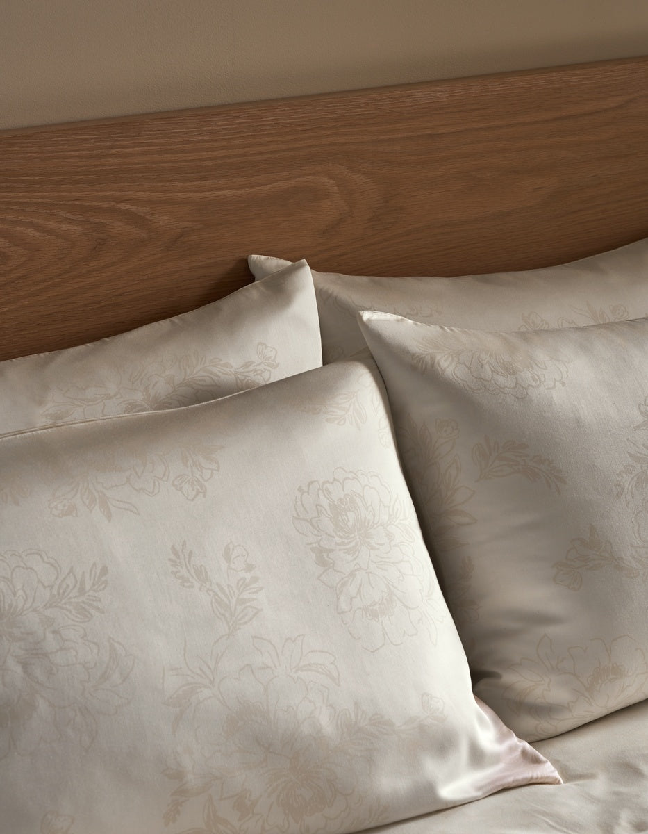Close-up of cream-colored damask pillowcases with a subtle floral pattern, arranged on a bed with a wooden headboard. 