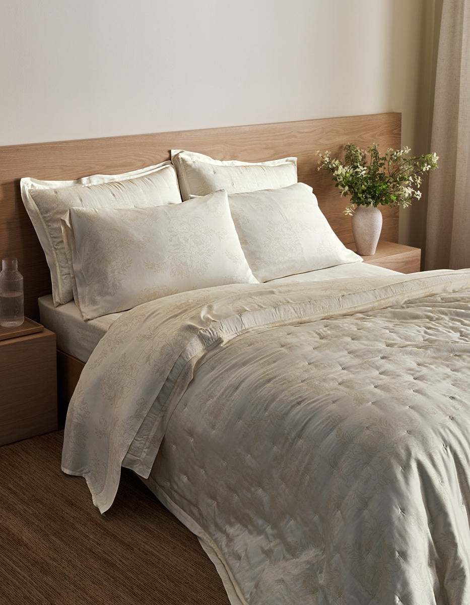 A neatly made bed featuring the Cozy Earth Bamboo Quilt and matching pillows in taupe sits beside a wooden nightstand with a white cup and modern lamp, set against a light wood-paneled wall.|Color:Walnut