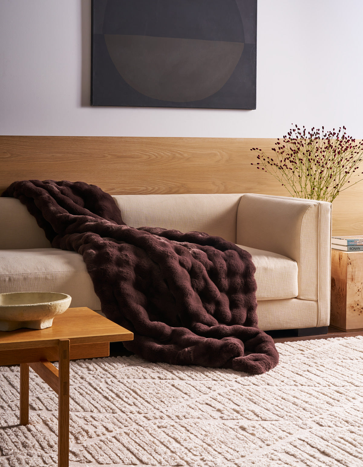 A modern living room featuring a cream-colored sofa, adorned with the Cozy Earth Bubble Cuddle Blanket, complemented by a dark purple textured blanket, a wooden table, a patterned rug, and minimalist decor.|Color:Raisin