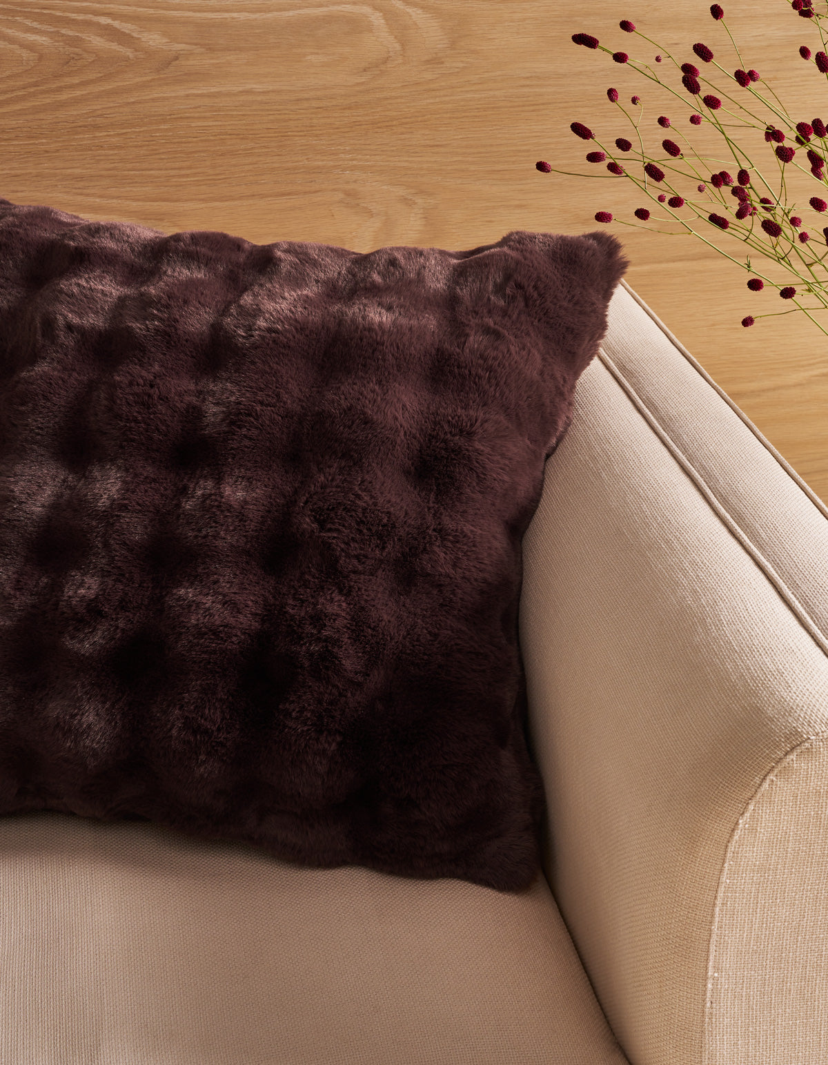A dark purple textured faux fur throw pillow resting on a beige fabric sofa, with a light wood floor and decorative dried stems in the background. 