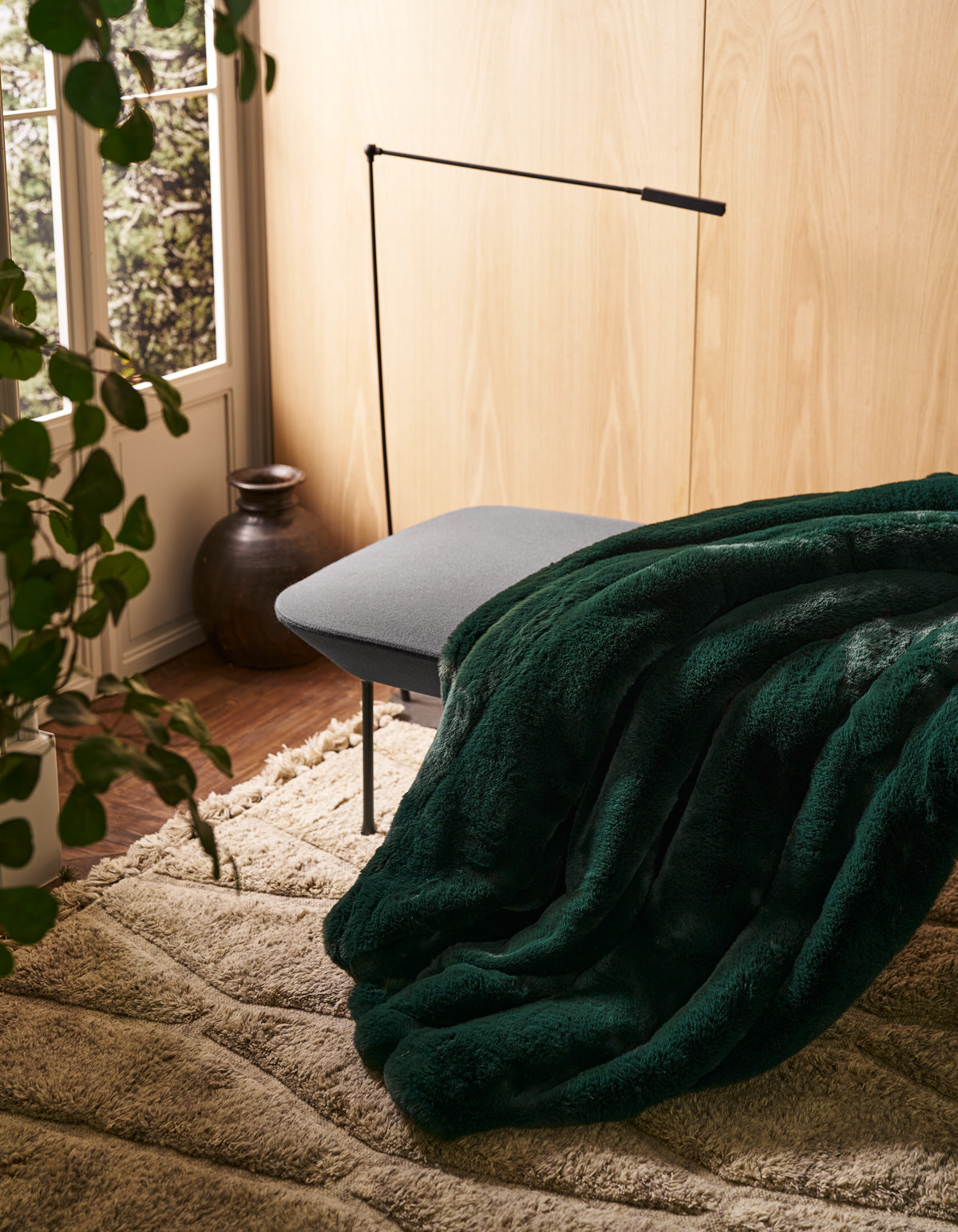 The Channel Cuddle Blanket by Cozy Earth, in a rich dark green faux fur, is draped over a gray bench in a cozy room featuring light wood walls, a beige rug, a potted plant, and natural light streaming through the window.