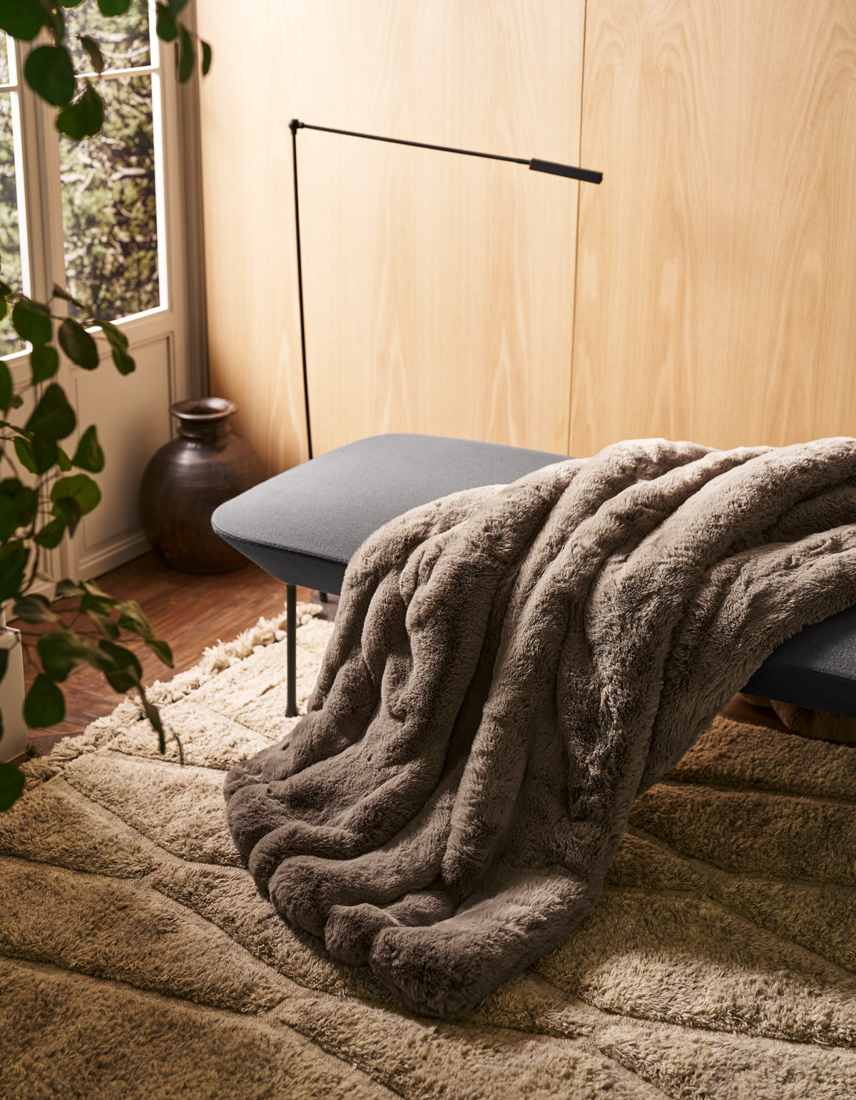 The Channel Cuddle Blanket from Cozy Earth, in a soft gray hue, is draped over a cushioned bench near a window. Nearby, a plant and a ceramic vase add natural charm as sunlight streams in, illuminating the textured beige rug and highlighting the warmth of the wooden wall.|Color:Walnut