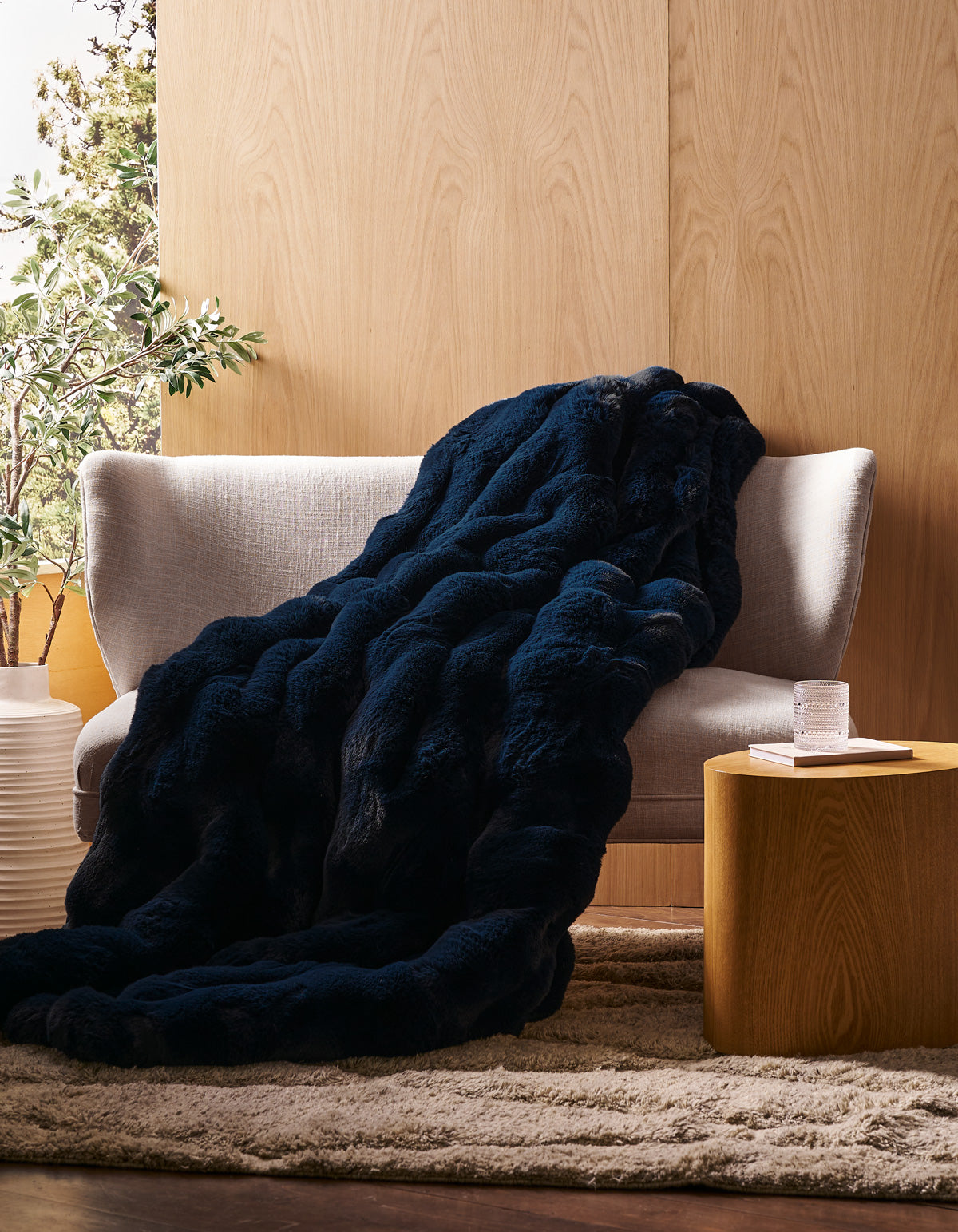 A Cozy Earth Double Bubble Cuddle Blanket in dark blue is draped over a light gray sofa, beside a round wooden side table holding a glass of water, all set in a cozy room with wood-paneled walls and a beige rug.|Color:Eclipse