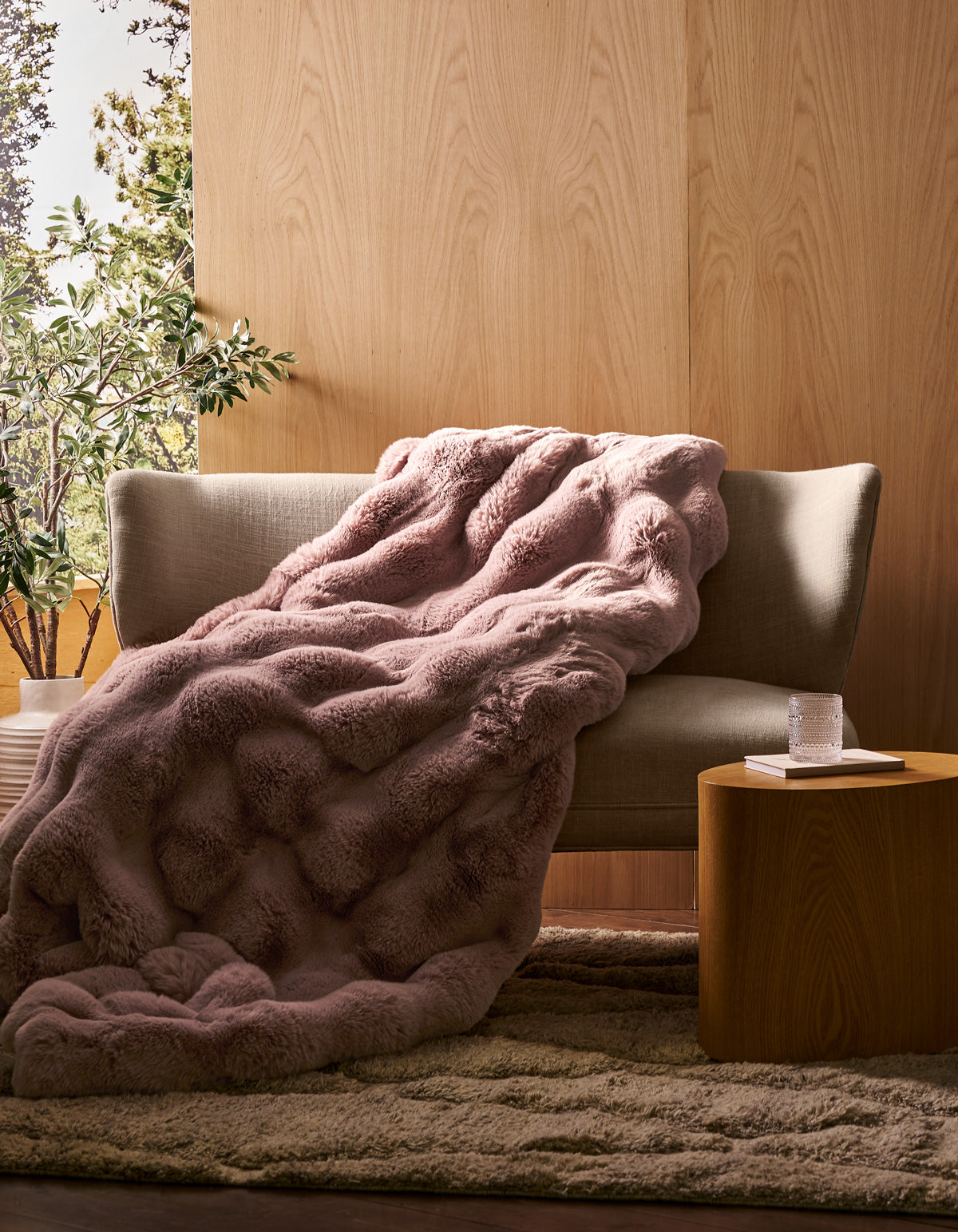 A beige sofa with the Cozy Earth Double Bubble Cuddle Blanket in textured pink draped over it sits beside a small wooden table holding a glass of water and a book, set in a cozy room with wood-paneled walls and a potted plant.|Color:Viola
