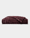 A folded, plush Bubble Cuddle Blanket by Cozy Earth in dark burgundy, featuring a soft, furry texture, displayed against a plain light gray background.|Color:Raisin