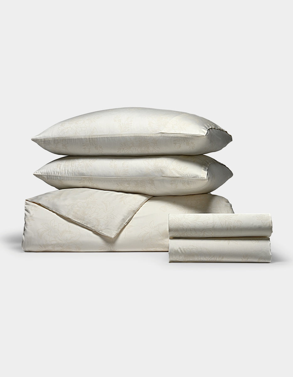 A Cozy Earth Bamboo Bedding Deluxe Bundle in cream, featuring a subtle floral pattern, with two pillows stacked atop a folded duvet and neatly folded sheets, displayed on a plain white background.