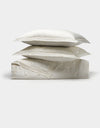 Two white Cozy Earth pillows are stacked atop a folded Bamboo Quilt Bundle, all crafted from a shiny, smooth fabric and set against a plain light background.|Color:Sonoma Floral in Vintage White