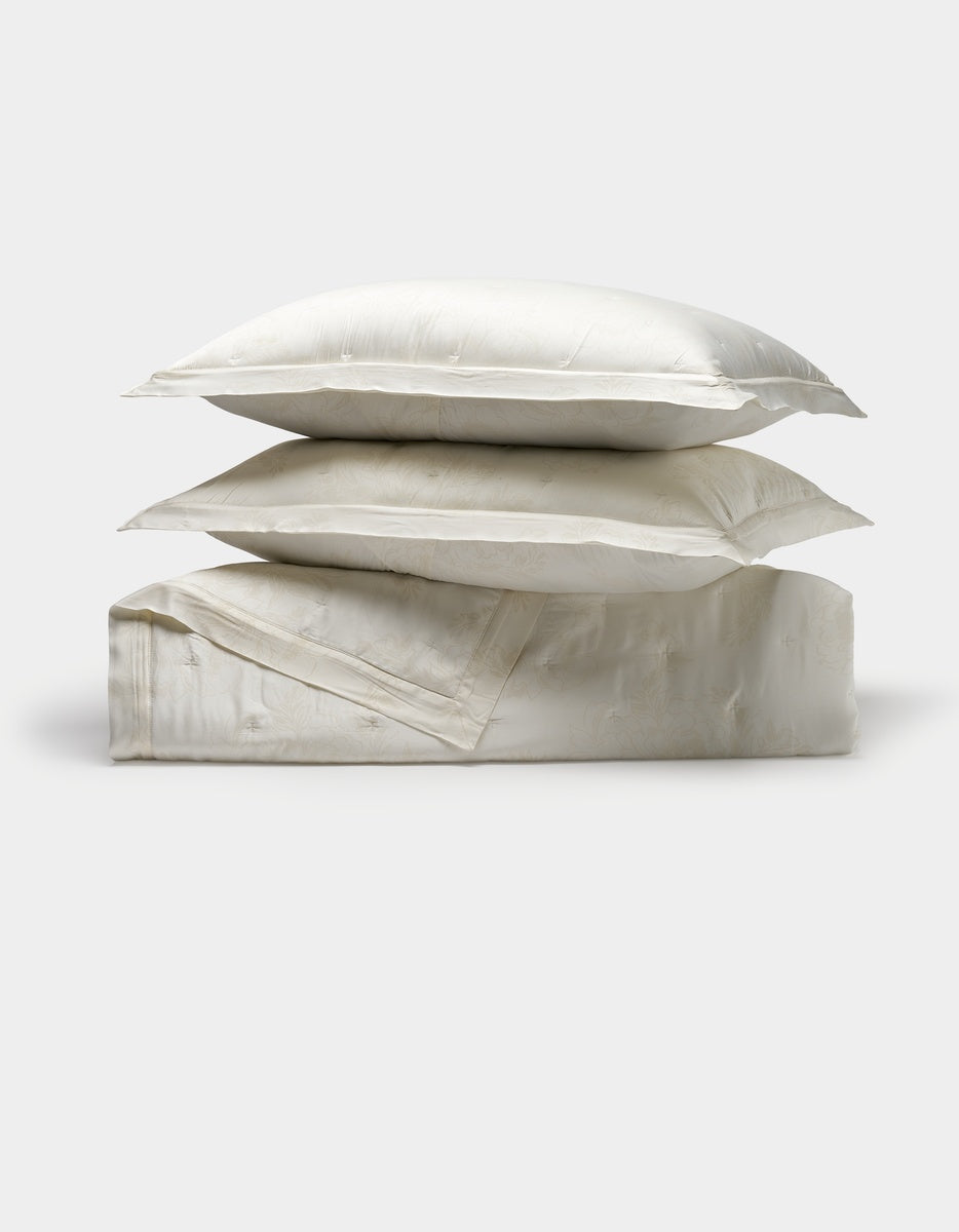 Two white Cozy Earth pillows are stacked atop a folded Bamboo Quilt Bundle, all crafted from a shiny, smooth fabric and set against a plain light background.