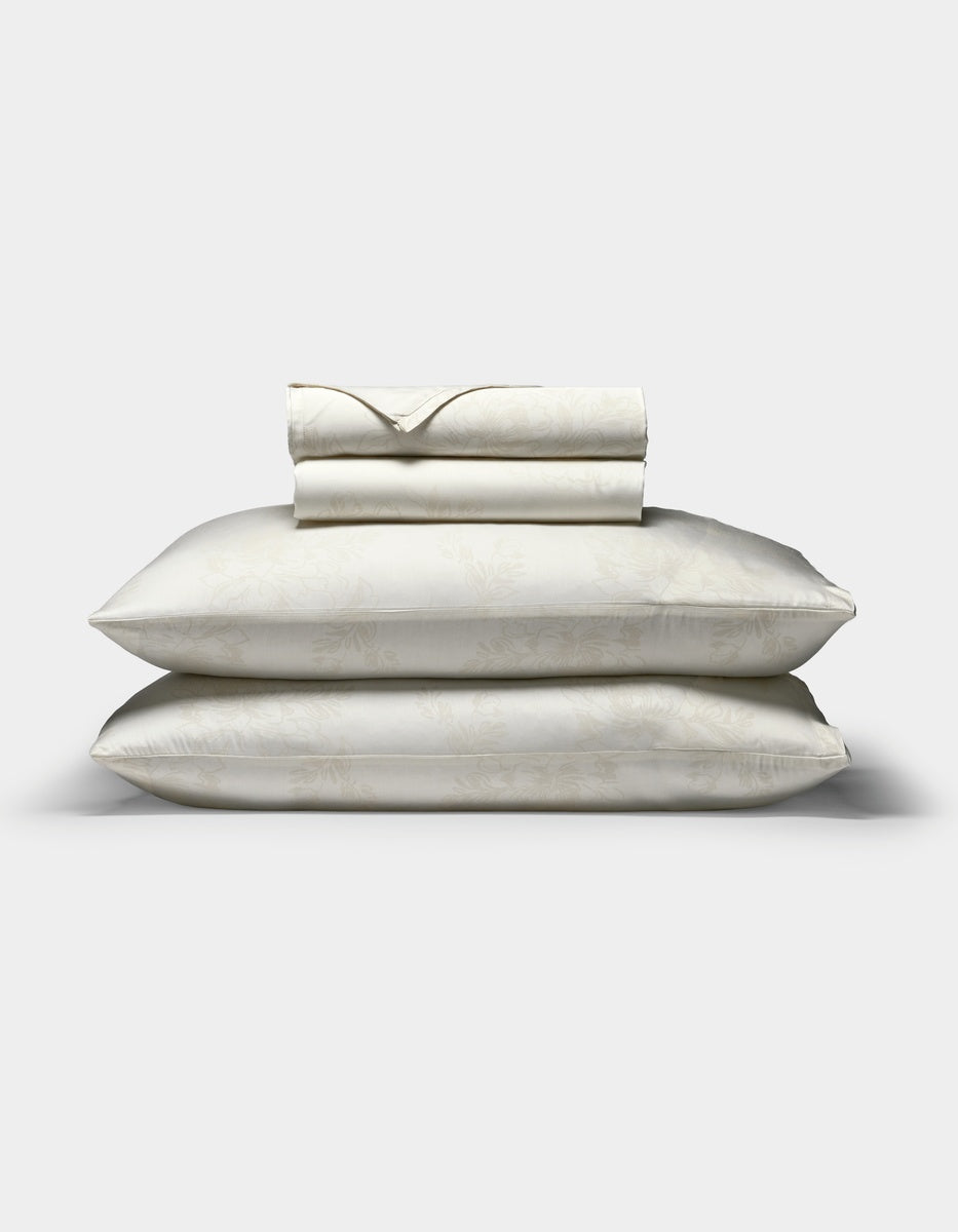 A Cozy Earth Bamboo Sheet Set in cream, featuring a subtle floral pattern, with two pillows in matching pillowcases stacked beneath neatly folded sheets, displayed on a plain white background.