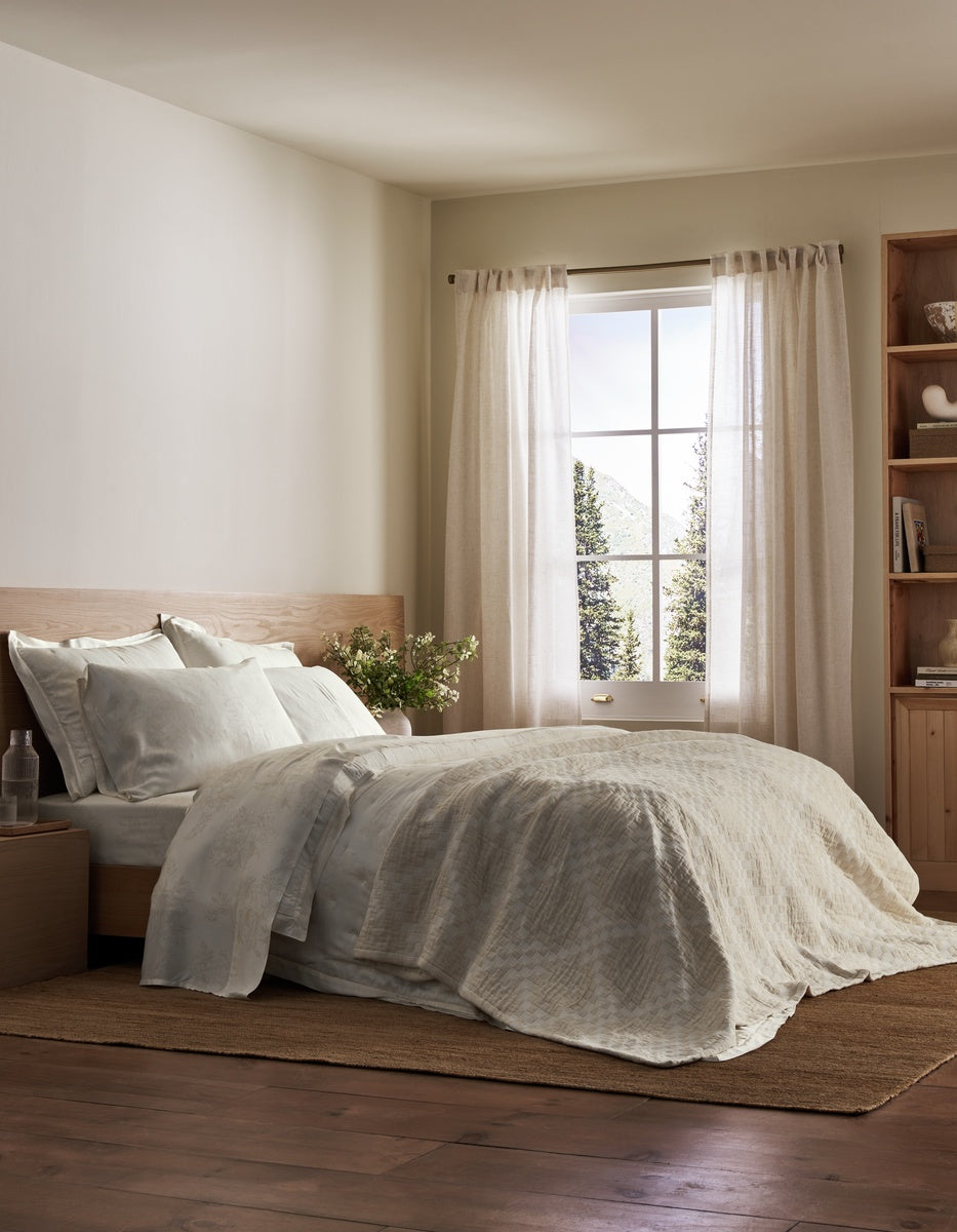 A cozy bedroom featuring a neatly made bed dressed in the Sonoma Stitch Quilt by Cozy Earth, complemented by white bedding, light curtains, a bedside table with a glass and flowers, a bookshelf, and sunlight streaming through a window that frames views of trees and mountains outside.
