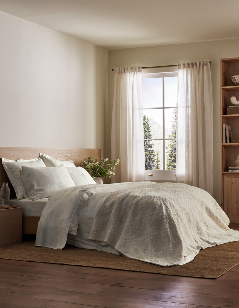 A cozy bedroom featuring a neatly made bed dressed in the Sonoma Stitch Quilt by Cozy Earth, complemented by white bedding, light curtains, a bedside table with a glass and flowers, a bookshelf, and sunlight streaming through a window that frames views of trees and mountains outside.