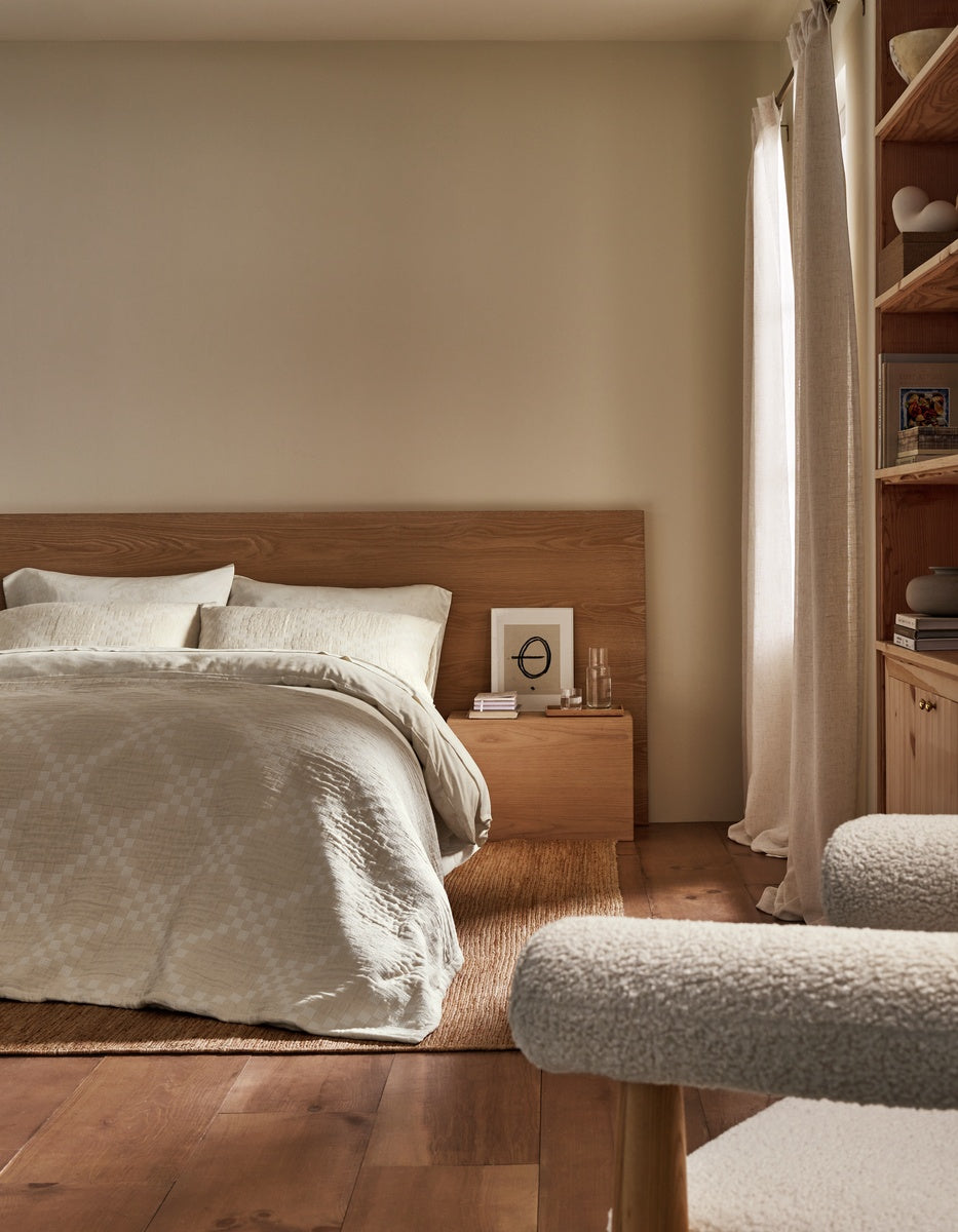 A cozy, minimalist bedroom featuring a wooden bed dressed in the Sonoma Stitch Quilt by Cozy Earth, light bedding, a wooden nightstand adorned with books and glasses, soft light filtering through curtains, and built-in shelves on the right.
