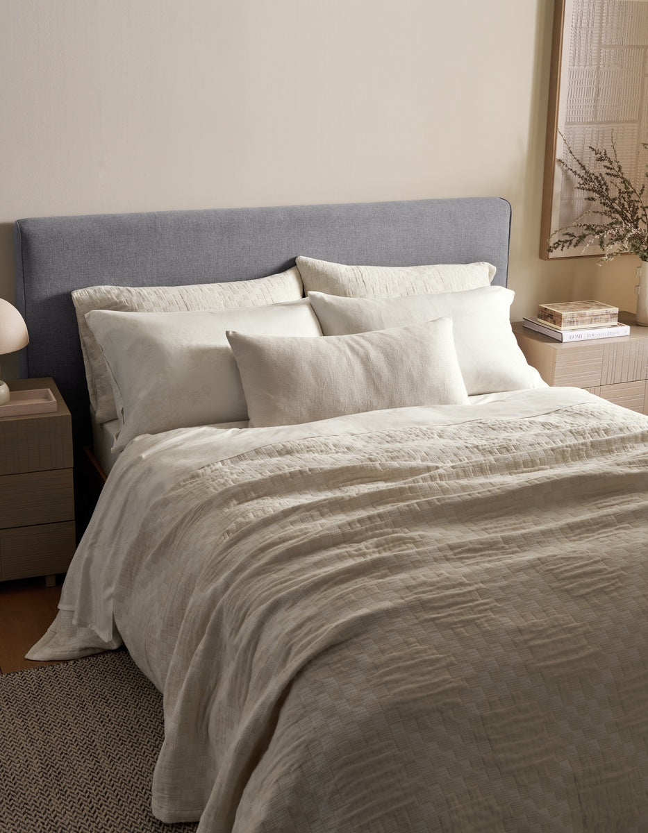 A neatly made bed features a gray upholstered headboard, cream-colored bedding, multiple pillows including the Sonoma Throw Pillow by Cozy Earth, and a beige quilt. Beside the bed, a nightstand holds a lamp and books, while decorative accents adorn the walls and tabletop.
