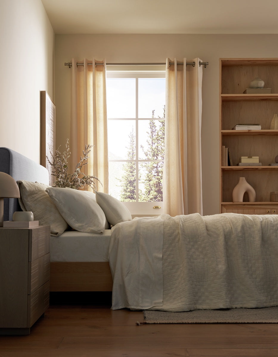 A sunlit bedroom features a neatly made bed adorned with light beige bedding and a Cozy Earth Sonoma Throw Pillow. A wooden nightstand sits nearby, while shelves decorated with tasteful accents and large windows with sheer curtains let in abundant natural light.