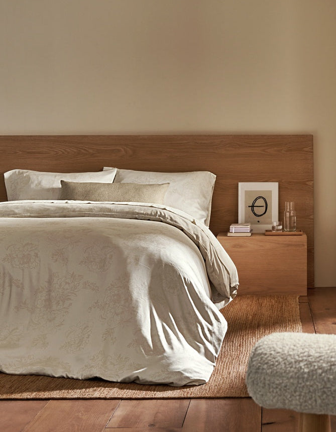 A cozy bedroom featuring a wooden bed with light bedding and accented by the Cozy Earth Sonoma Throw Pillow. A nightstand holds books and a glass of water, while sunlight streams through beige curtains on a large window. Wooden floors and shelves enhance the room’s warmth.