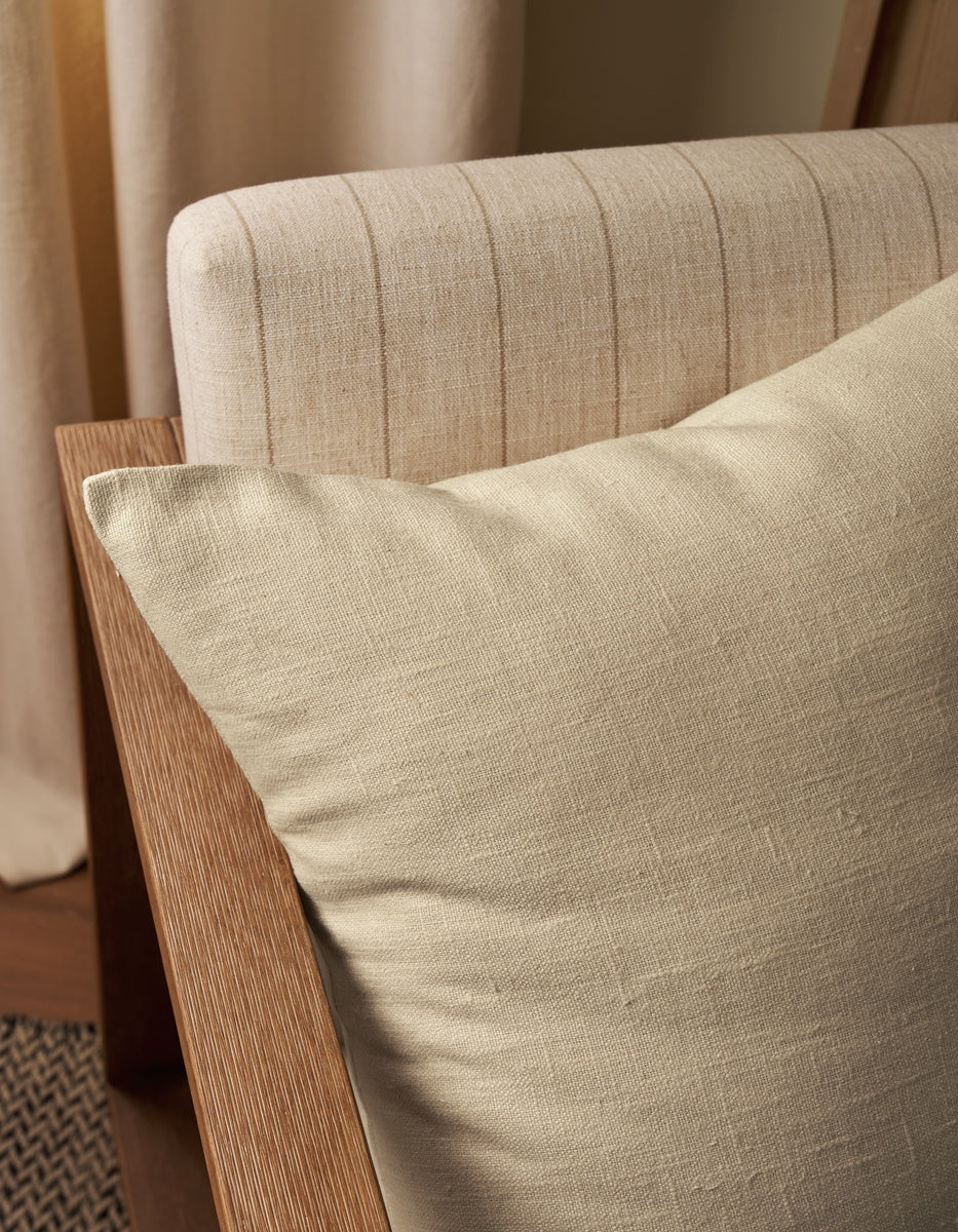 A close-up of the Sonoma Throw Pillow by Cozy Earth, in beige, resting on a light wood-framed chair with a cream, vertically-striped upholstered backrest. Neutral curtains and a woven rug are partially visible in the background.
