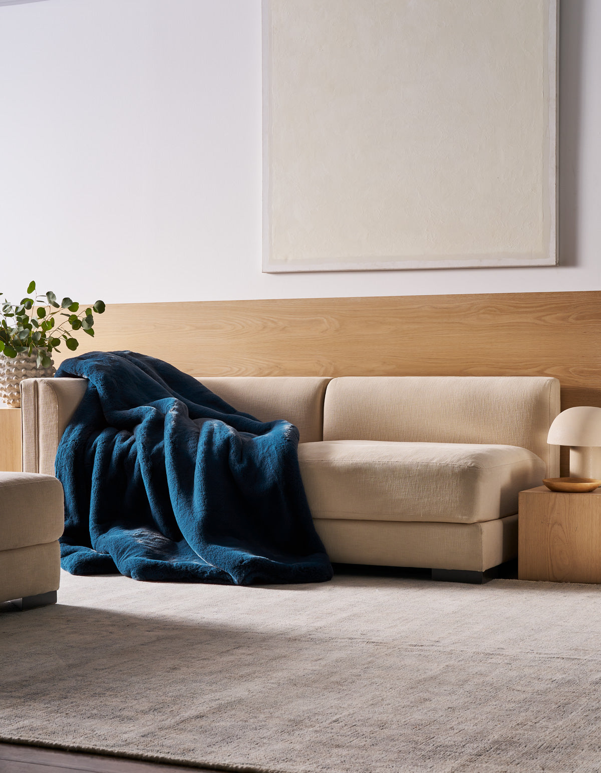 A beige sectional sofa is draped with the Cozy Earth Superplush Cuddle Blanket in deep blue, positioned beside a small wooden side table topped with a lamp and a plant. The space features a light rug, wood paneling, and a large blank canvas displayed on the wall.|Color:Deep Water