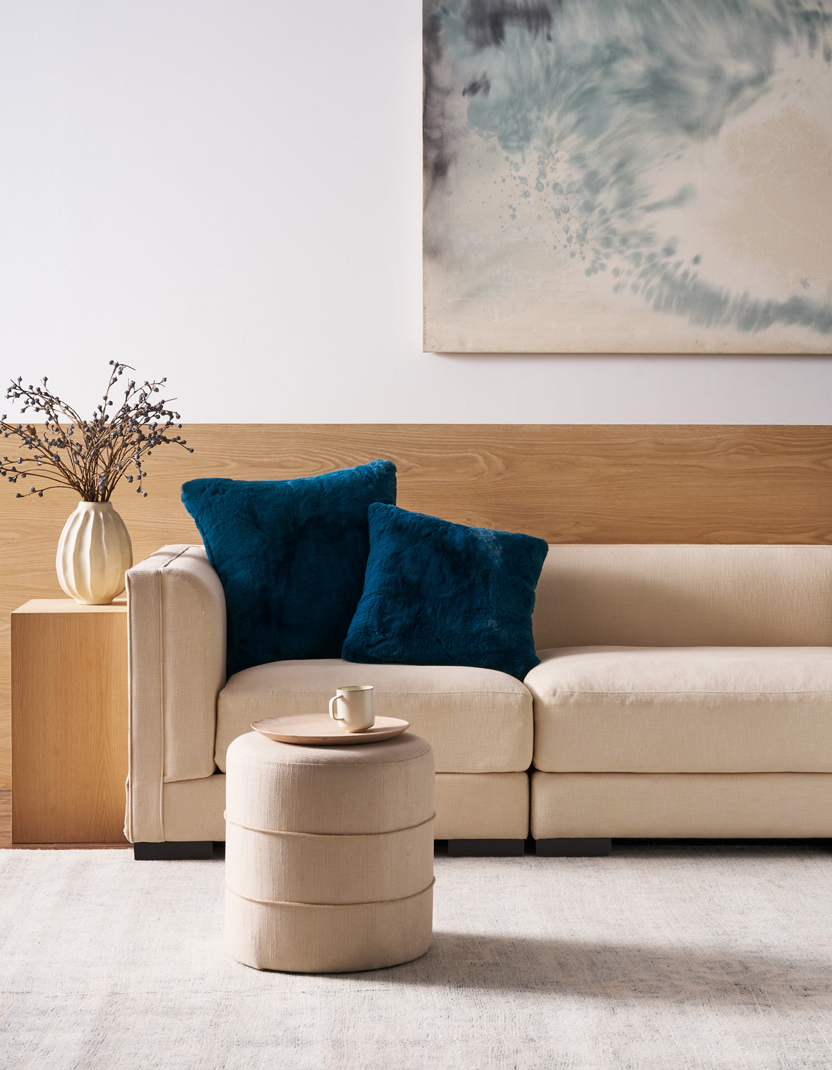 A modern living room scene featuring a beige sectional sofa with two plush dark teal throw pillows, a matching cylindrical ottoman, a wooden side table with a vase of branches, and a large abstract painting on the wall. 