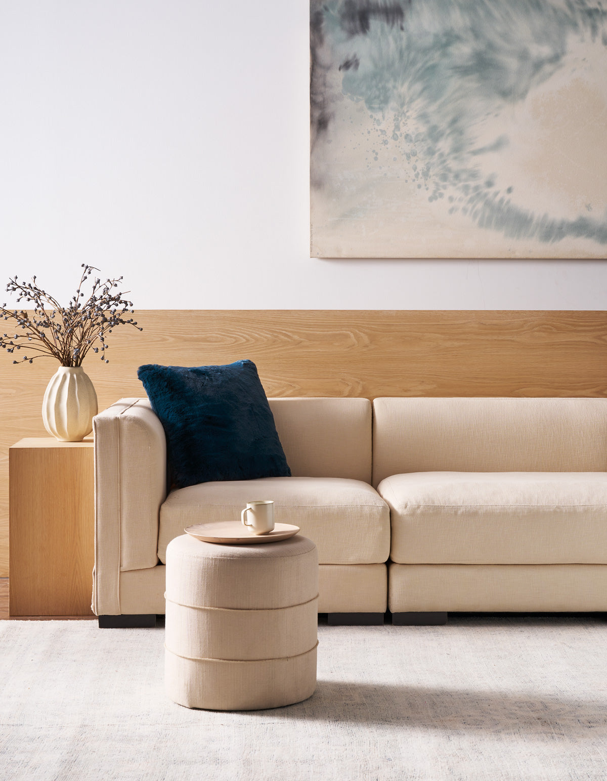 A modern living room featuring a beige sofa, the Superplush Cuddle Pillow by Cozy Earth in dark blue, a round ottoman holding a cup and saucer, a vase with dried branches, and abstract wall art in blue and gray tones displayed above wood paneling.|Color:Deep Water |Size:24" x 24"