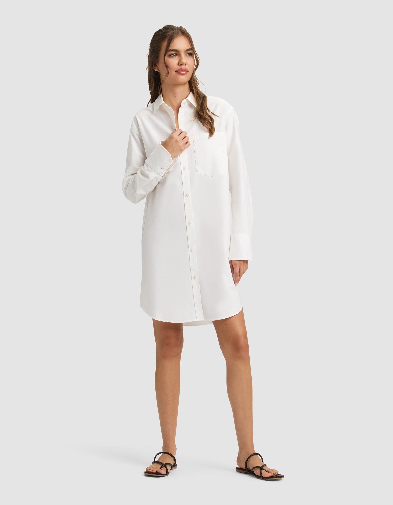 A woman with light brown hair stands against a plain background wearing the Cozy Earth Women's Camden Dress, a long-sleeved white button-up shirt dress with a front pocket, paired with black sandals. She gazes to the side with a relaxed expression. 