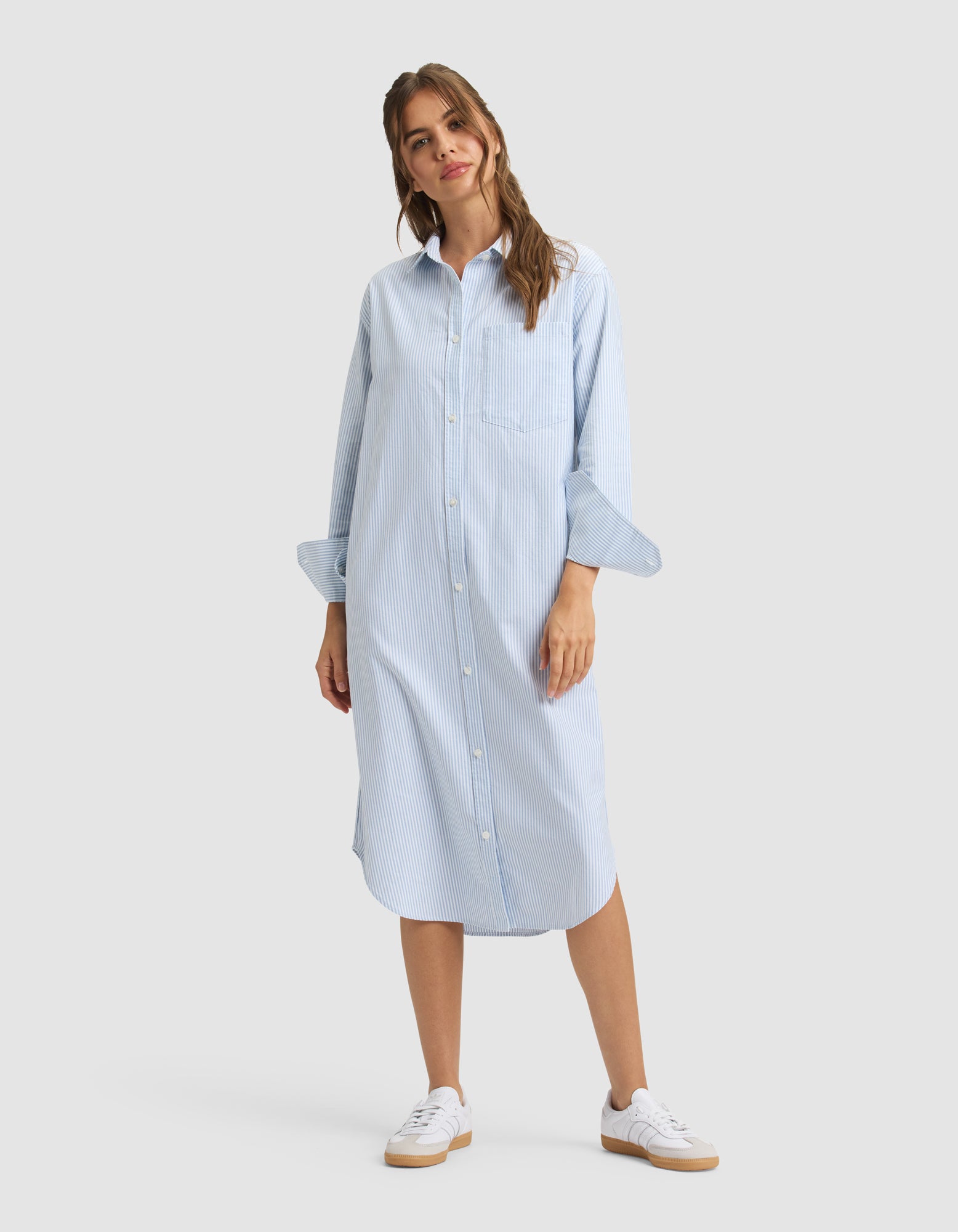 A woman wears the Cozy Earth Women's Camden Midi Dress, a loose light blue, long-sleeve, button-down shirt dress with subtle stripes. She pairs it with white sneakers and smiles slightly, posing with one hand by her side. 