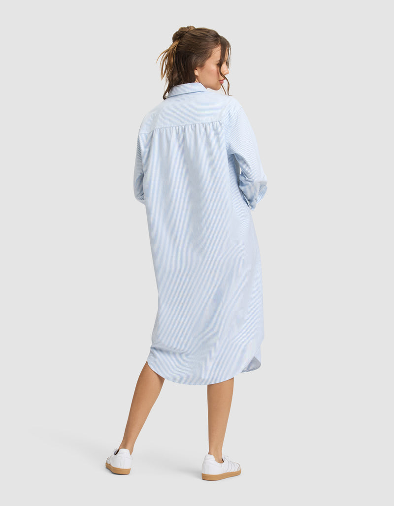 A woman faces away, wearing Cozy Earth's Women's Camden Midi Dress in light blue with subtle stripes and long sleeves, paired with white sneakers. Her hair is partially tied up, set against a plain light gray background.