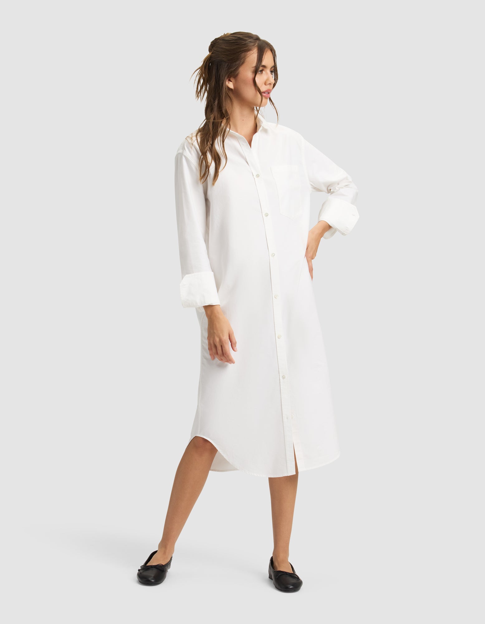 A woman wearing the Cozy Earth Women's Camden Midi Dress with rolled-up sleeves and black flats smiles, looking to the side with wavy brown hair styled half-up and one hand on her hip. 