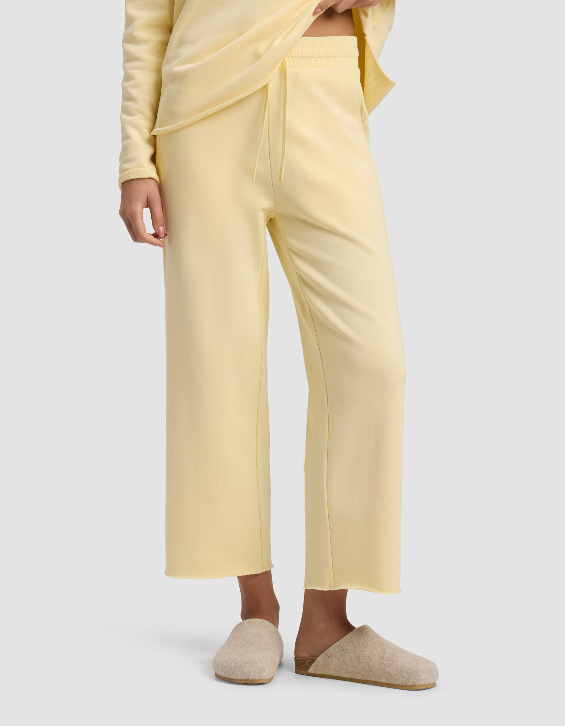 A person is wearing Cozy Earth's Women's Chelsea Cropped Pant in light yellow with an elastic waistband and drawstring, paired with a matching top and beige slippers, standing against a plain background.|Color:Meringue