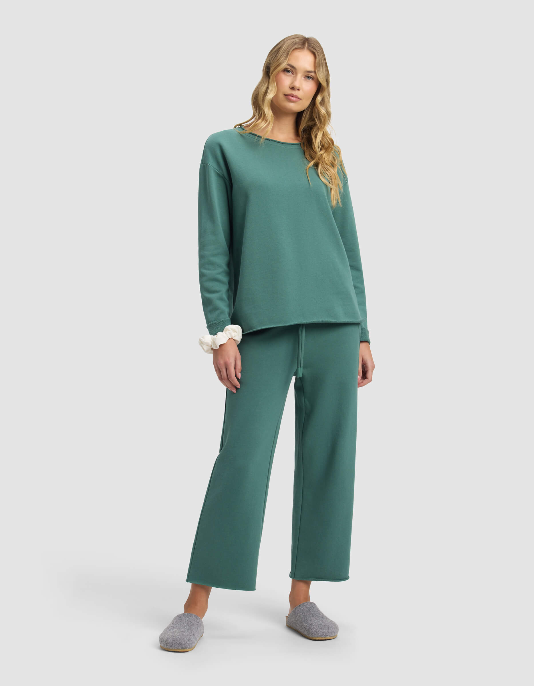 A woman with long blonde hair wears Cozy Earth’s Women’s Chelsea Cropped Pant in teal with a matching relaxed sweatshirt, a white scrunchie on her wrist, and gray slippers, standing against a plain light gray background.