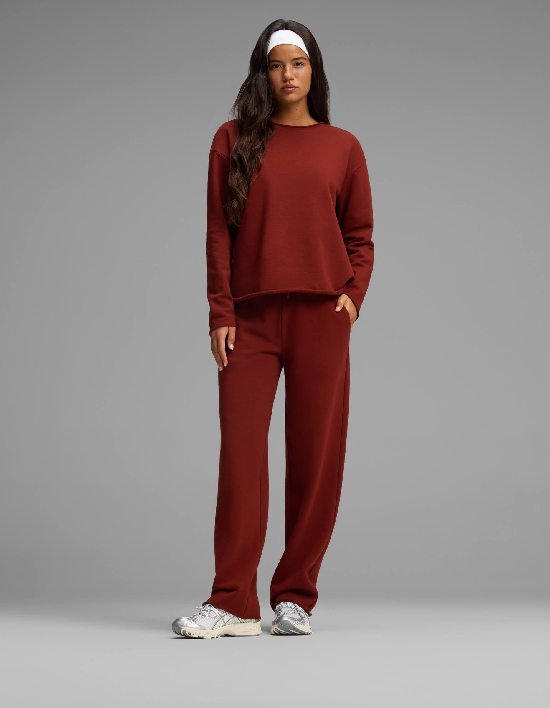 A woman stands against a plain gray background wearing the Cozy Earth Women's Chelsea Set—a matching rust-colored long-sleeve top and pants. She has long dark hair, a white headband, and white sneakers, with one hand in her pocket.|Color:Currant