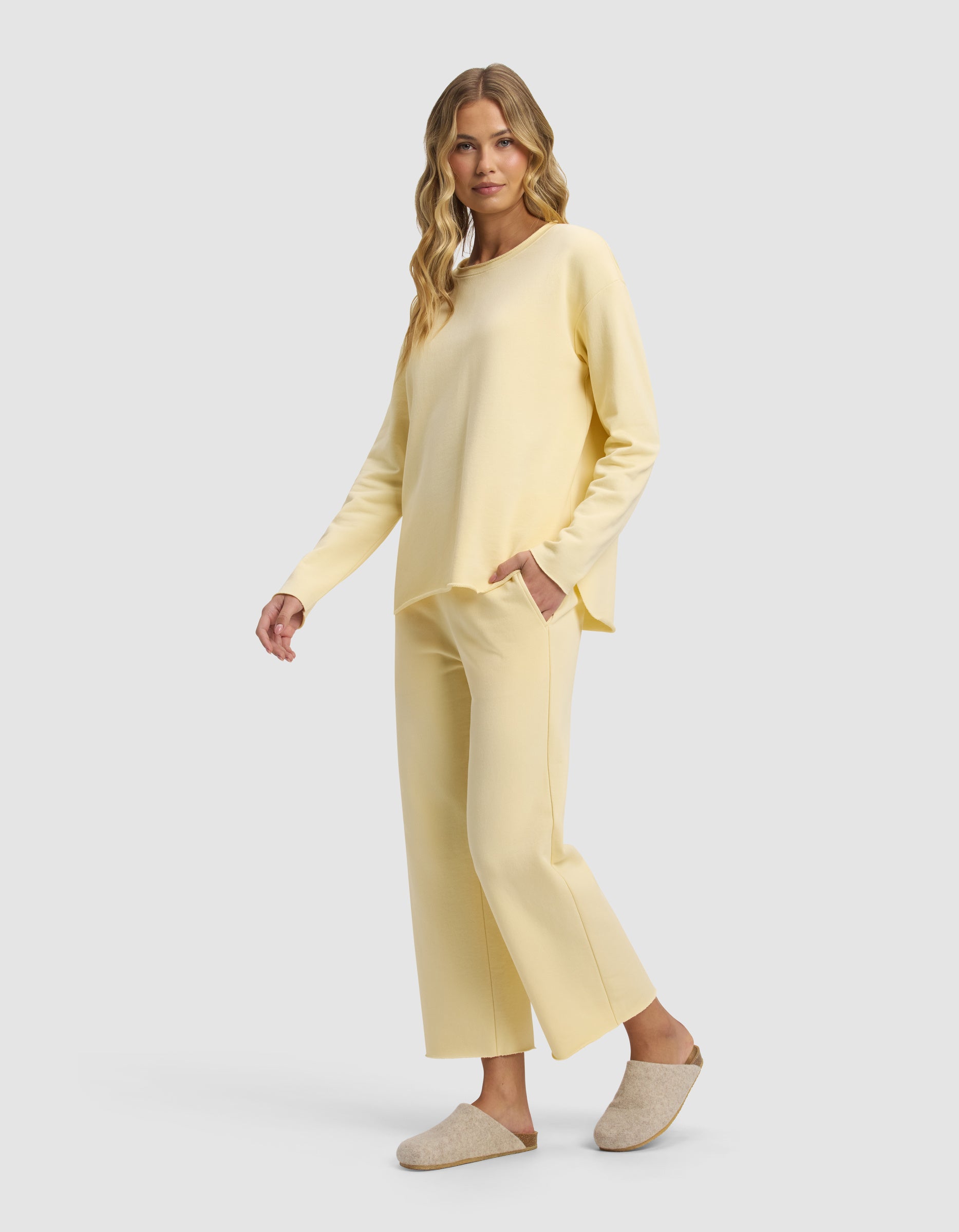 A woman with long wavy hair models the Cozy Earth Women's Chelsea Cropped Set in light yellow, featuring long sleeves and wide-leg pants. She stands sideways with one hand in her pocket, wearing beige slippers against a plain light gray background.