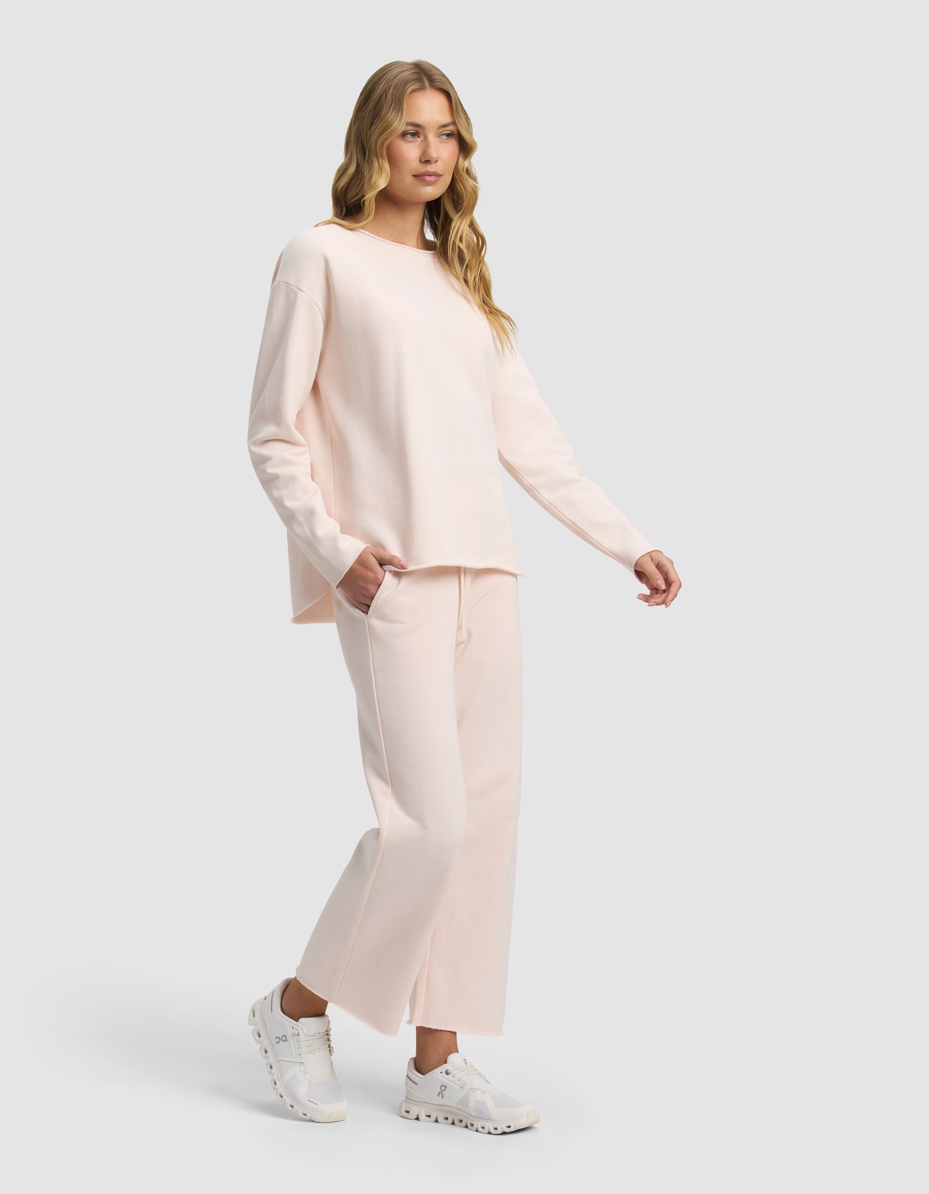 A woman with long blonde hair wears the Cozy Earth Women's Chelsea Cropped Set in light pink, paired with white sneakers, standing against a plain light gray background.