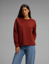 A woman with long dark hair wears the Cozy Earth Women's Chelsea Pullover in a rust color paired with blue jeans, standing against a plain gray background with a neutral expression.|Color:Currant
