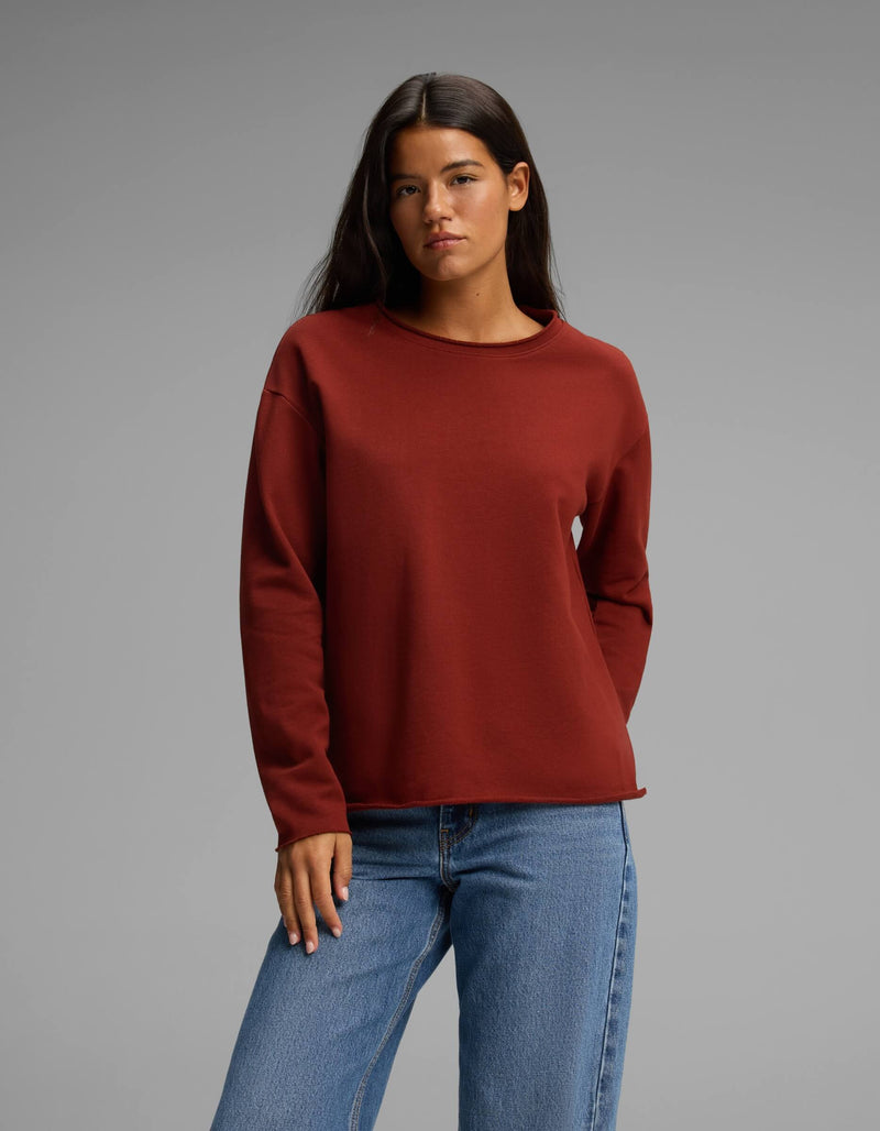 A woman with long dark hair wears the Cozy Earth Women's Chelsea Pullover in a rust color paired with blue jeans, standing against a plain gray background with a neutral expression.