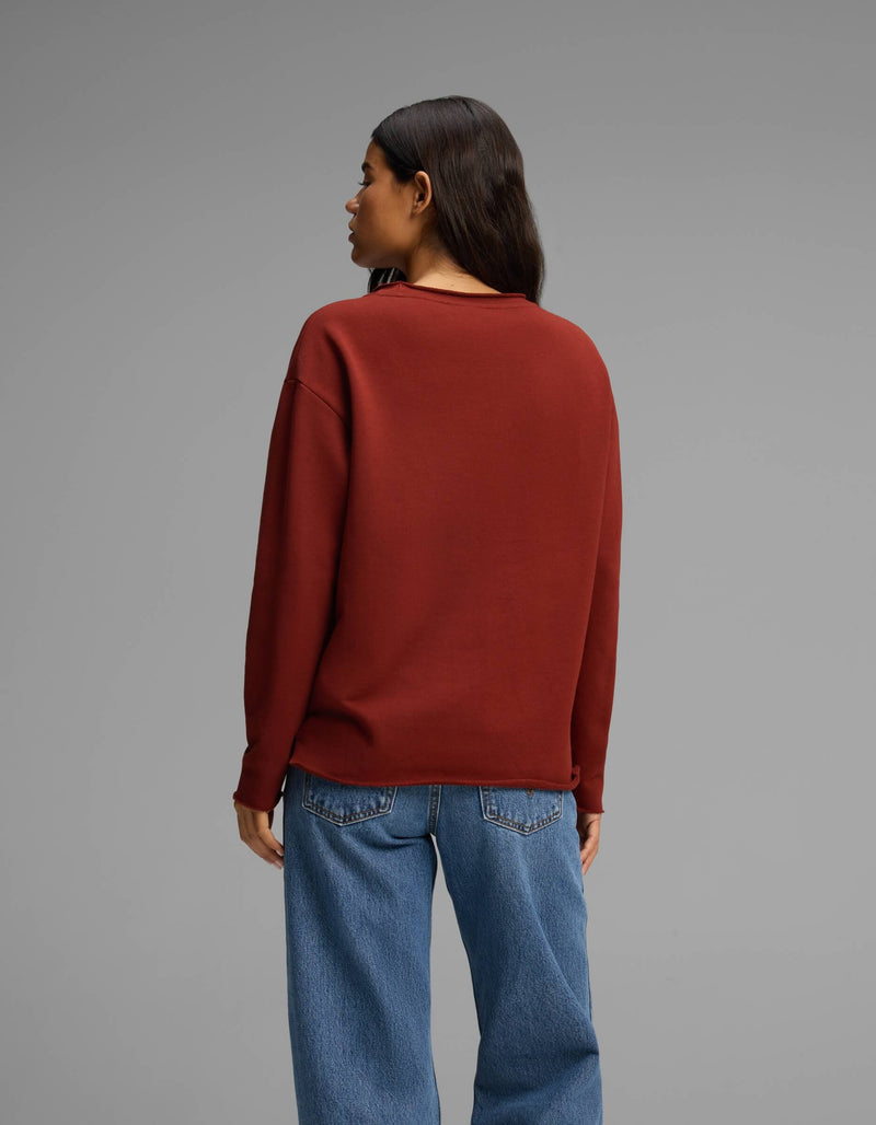 A woman with long dark hair, wearing the Cozy Earth Women's Chelsea Pullover in maroon and blue jeans, stands facing away from the camera against a plain gray background.