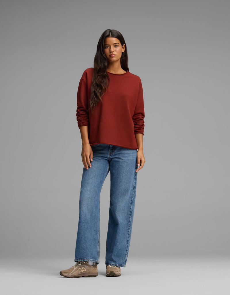 A woman with long dark hair wears the Cozy Earth Women's Chelsea Pullover in a rust shade, paired with blue jeans and brown sneakers. She stands against a plain gray background, looking at the camera with a relaxed posture.