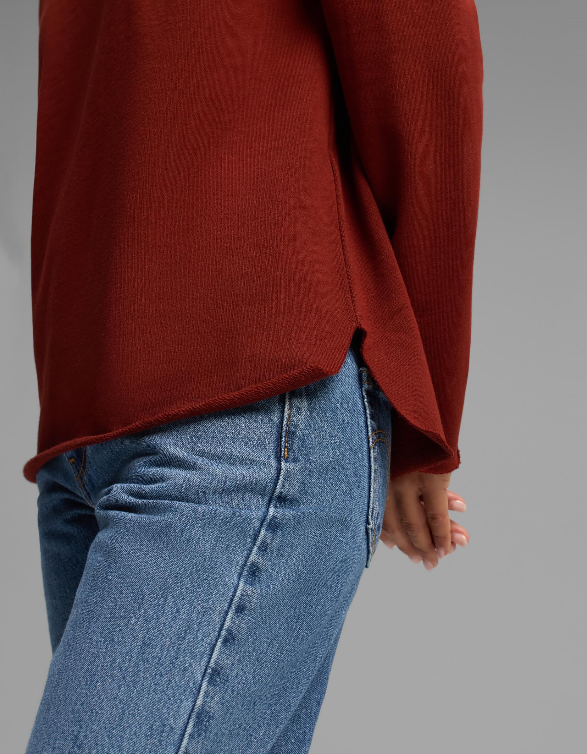 A person shown from the waist down, standing sideways with one hand behind their back against a plain gray background, wearing blue jeans and the Women's Chelsea Pullover in rust red by Cozy Earth.