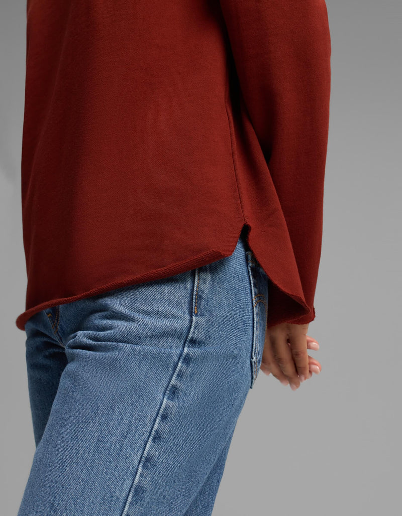 A person shown from the waist down, standing sideways with one hand behind their back against a plain gray background, wearing blue jeans and the Women's Chelsea Pullover in rust red by Cozy Earth.