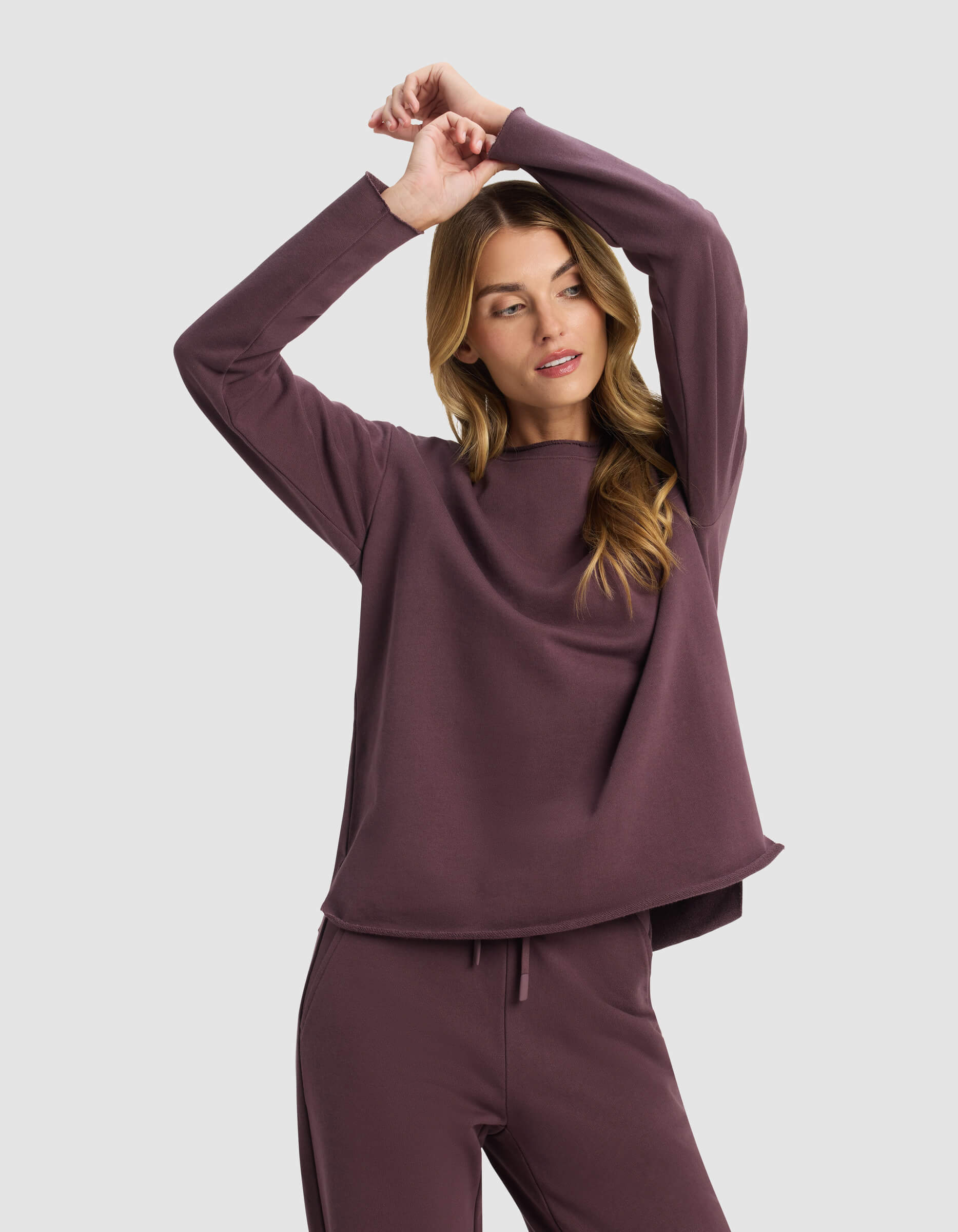 A woman models the Cozy Earth Women's Chelsea Pullover in mauve, paired with matching pants, standing against a light gray background with her arms raised and crossed above her head, gazing to the side.|Color:Fig