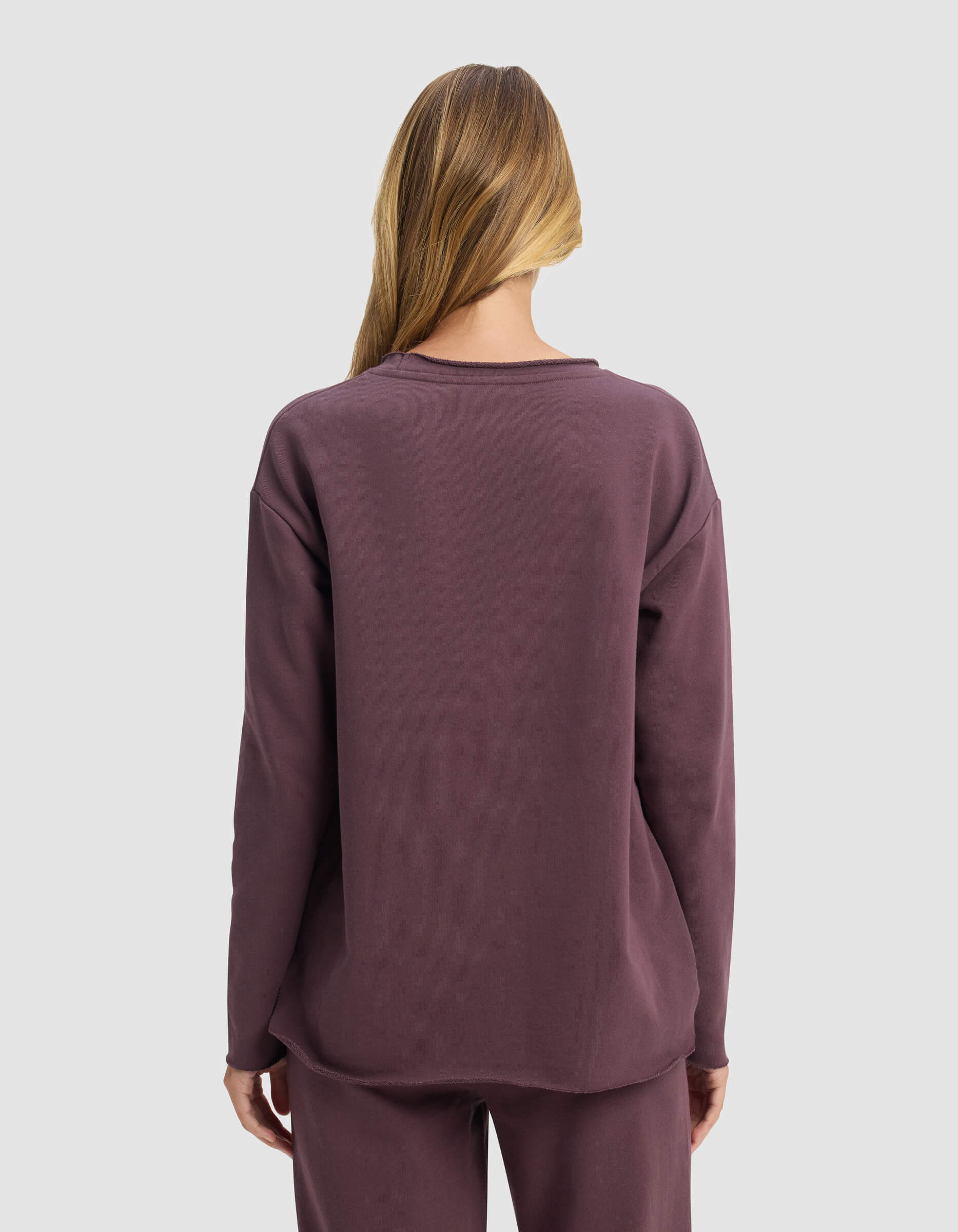 A person with long, light brown hair faces away, wearing the Cozy Earth Women's Chelsea Pullover and matching pants in mauve against a plain light gray background.