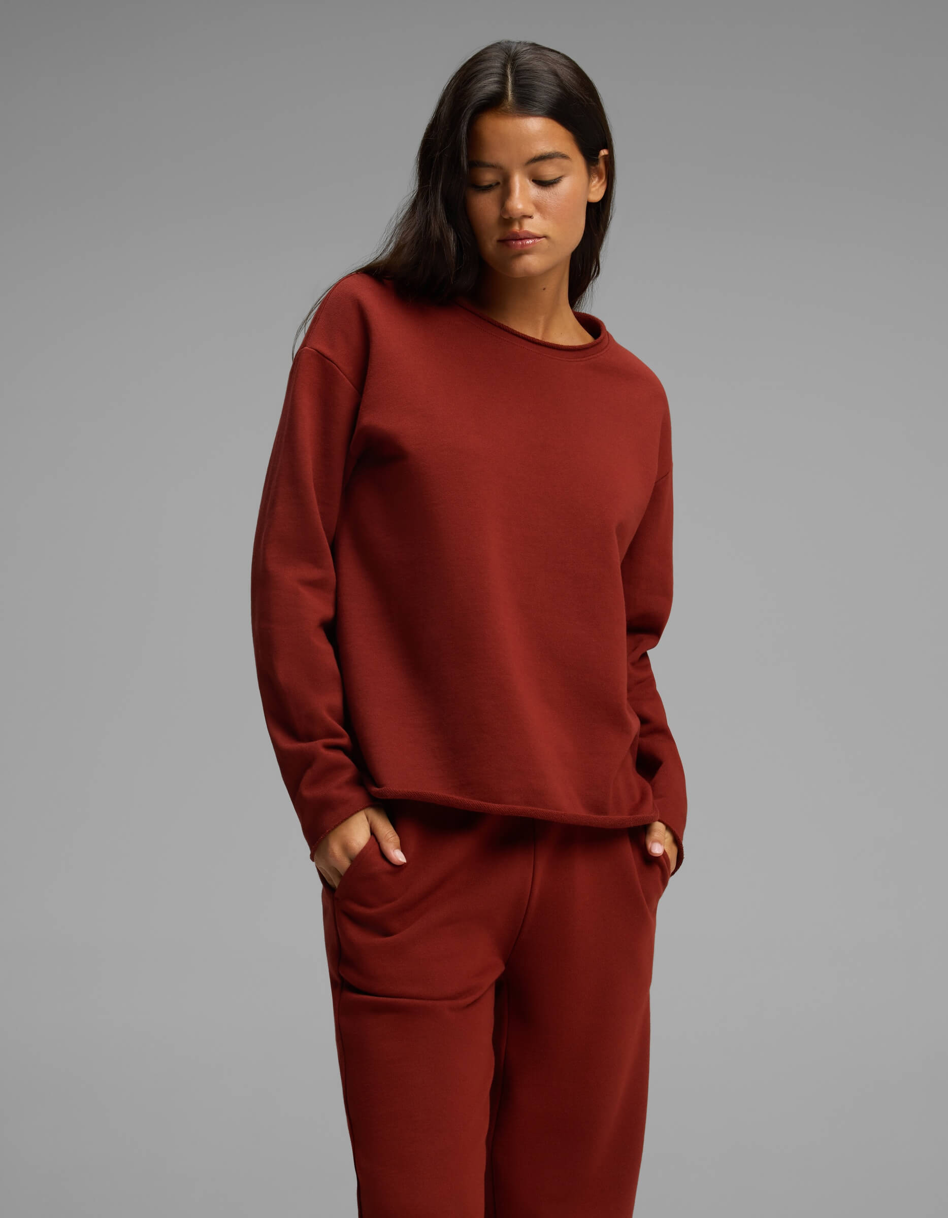 A person stands against a gray background wearing the matching maroon Women's Chelsea Set by Cozy Earth, with hands in pockets and looking down with a neutral expression.